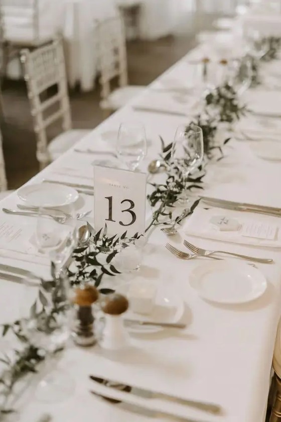 an ethereal neutral wedding table setting with white linens and chargers, a greenery runner, some candles seems simple but very cool