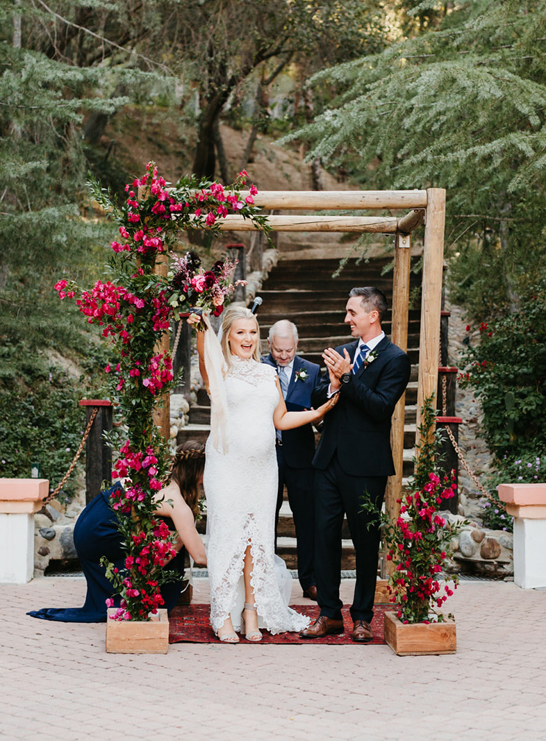 The groom was wearing a black suit with a navy tie and brown shoes, and the wedding arch was decorated with bougainvilleas