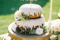 a two-tier bundt wedding cake with creamy drip, white blooms and greenery and a gold calligraphy topper