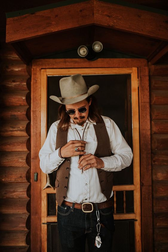 a boho western groom's look with a white shirt, navy jeans, a brown waistcoat, a brown belt and a grey hat, some western accessories