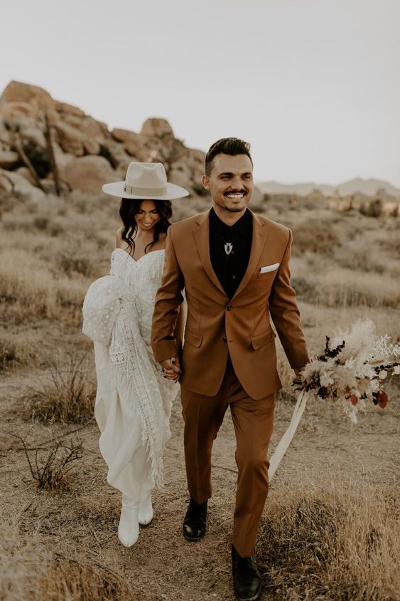 a bold boho groom's look with a black shirt, a rust pantsuit, a bolo tie and black shoes is a catchy and cool idea for a wedding