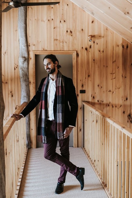 a casual and comfy winter groom's look with a black velvet blazer, burgundy pants, a white button down and a bright scarf
