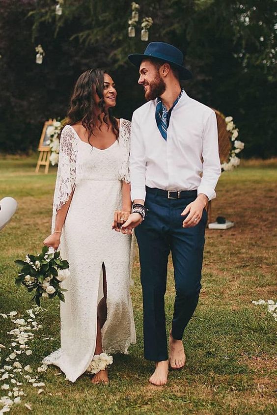 a relaxed groom's outfit with a white shirt, navy trousers, a navy neck tie and a hat for a spring or summer boho wedding