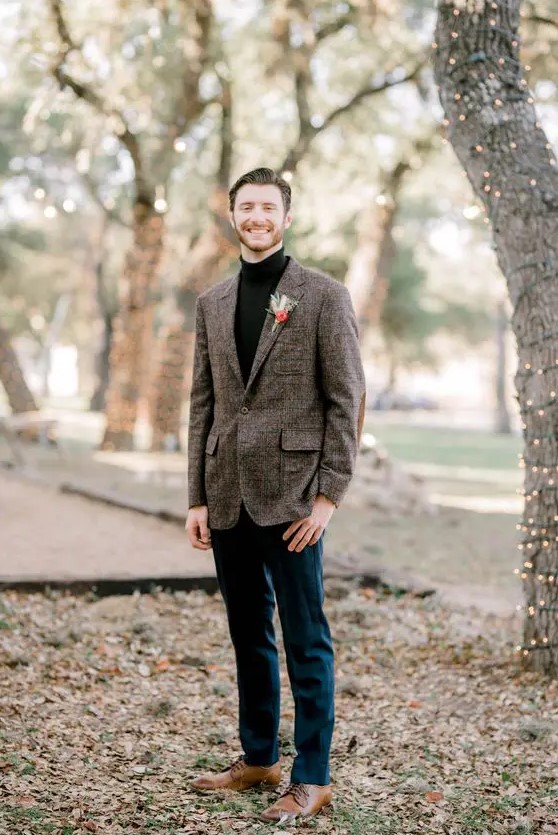 a simple boho winter groom look with a black turtleneck, a brown plaid blazer, navy pants, brown shoes and a bold boutonniere