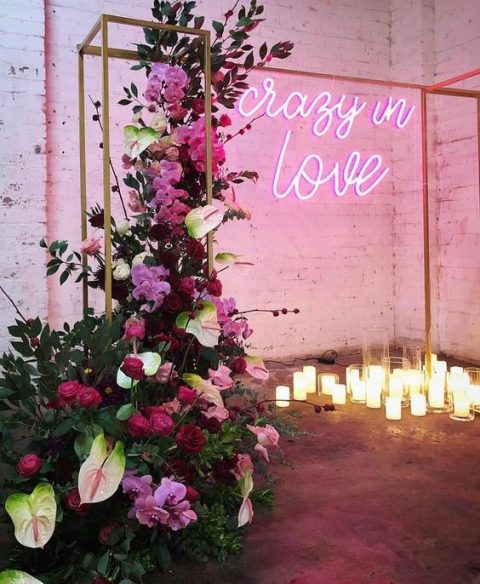 a beautiful wedding ceremony space with candles, an arch, lush florals in red, burgundy and pink and a pink neon sign