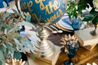 a celestial wedding table with a blue and gold globe, greenery, books, blue glasses and gold cutlery