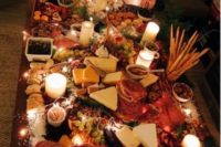 a delicious grazing table with various types of cheese, charcuterie, bread and crackers, some fruit and olives and candles and lights
