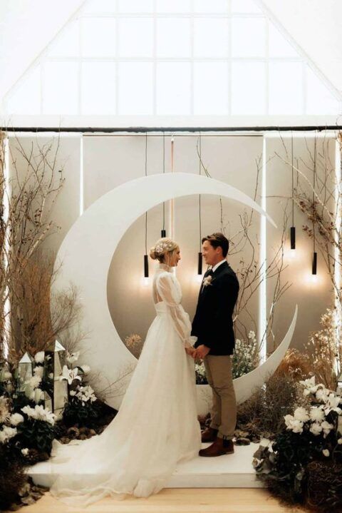 a modern celestial wedding backdrop of a half moon, white blooms, mirror pieces, pendant lamps and branches