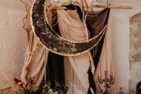 a moody celestial wedding backdrop with blush and black fabric, a black half moon, dark blooms and leaves and dried branches