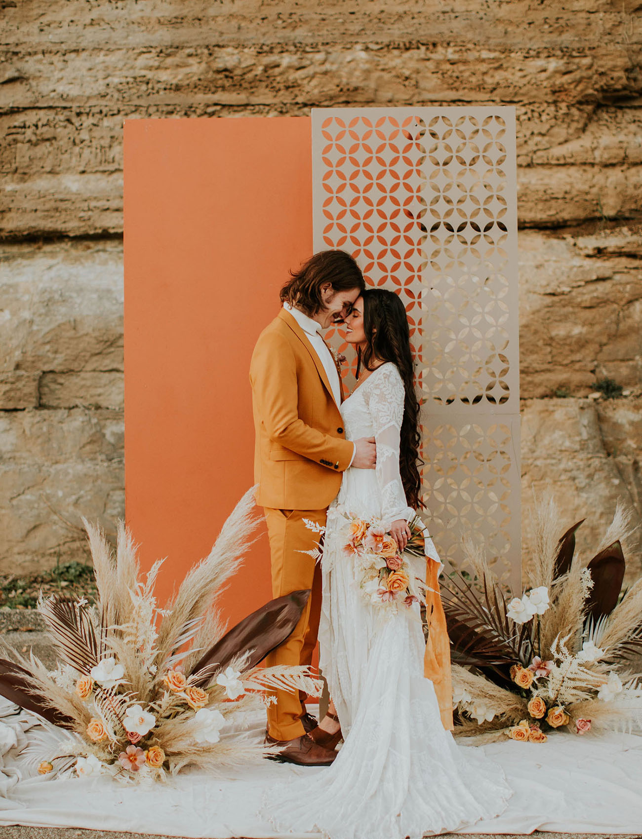 A creative 70s inspired wedding backdrop of an orange and a neutrla laser cut screen, dried fronds, leaves and blooms