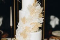 A simple white three tiered cake with beige and light brown feathers one the wooden stand