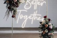 a chic wedding arch of metal, with lush blooms and greenery and a neon sign for a modern wedding