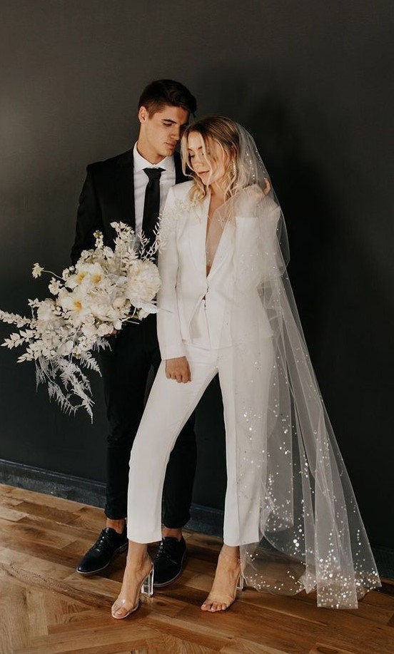 a chic white pantsuit with cropped pants, sheer heels and a long veil with sequins for a comfy reception look