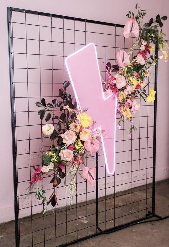 a funky wedding backdrop of mesh, a pink neon sign, pink and yellow blooms and greenery and dark leaves