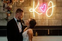 a pretty wedding arch with peachy, rust and neutal blooms and a neon sign with monograms and a heart is all cool