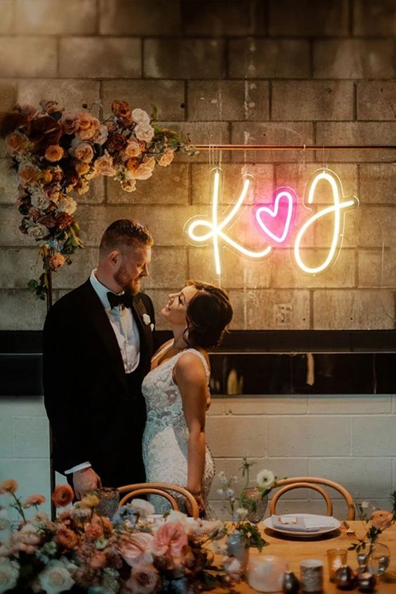 a pretty wedding arch with peachy, rust and neutal blooms and a neon sign with monograms and a heart is all cool