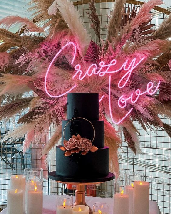a wedding cake backdrop with a lot of pampas grass, dried leaves, a pink neon sign, a black wedding cake surrounded with pillar candles