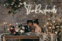 a wedding sweetheart table with an overhead floral installation, a neon sign, floral arrangements and black candles
