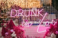 an outdoor wedding lounge with a pink seat, bold pink baby’s breath and fronds and a pink neon sign in the backdrop