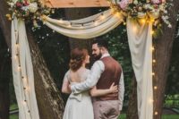 07 a cozy rustic wedding arch with string lights, neutral fabric, lush florals and candles on tree slices around