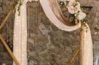 08 a glam hexagon wedding arch decorated with lush pastel and neutrla blooms, greenery, candles and a white and gold curtain with lights inside plus petals around