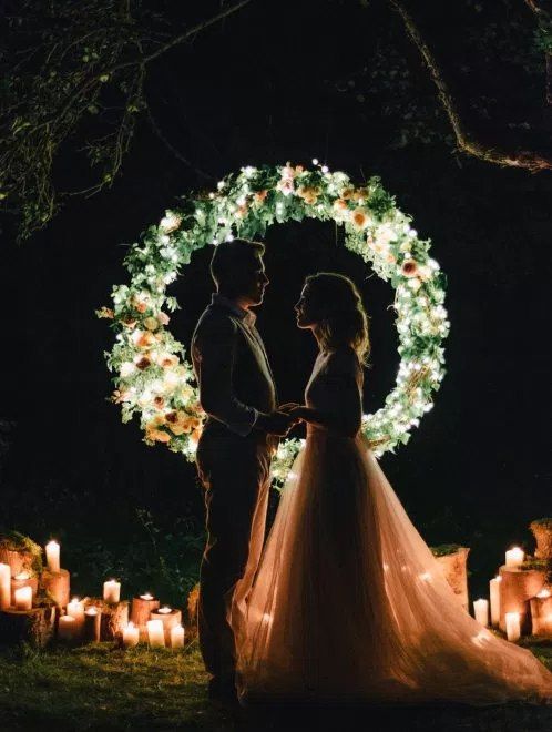 a mini round wedding arch fully made of greenery and blooms and lights inside it plus candles all around for a night ceremony