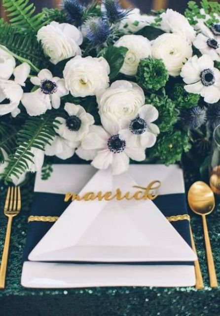 a bright and chic tablescape with lush greenery and white blooms, a navy and white napkin and a green sequin tablecloth plus gold cutlery