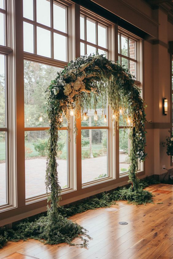 a rounded wedding arch fully covered with greenery and white blooms and some bulbs hanging down for an accent
