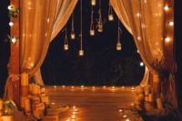 18 a simple and cool wedding arch with burlap curtains, string lights, candleholders hanging down and candles lining up the aisle