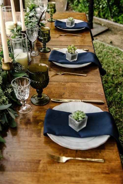 a relaxed wedding tablescape with pillar and tall candles, navy napkins, a eucalyptus table runner and blue and grey glasses