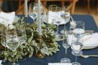 21 a simple and chic table setting with a navy tablecloth, a lush greenery runner, tall candles and grey plates