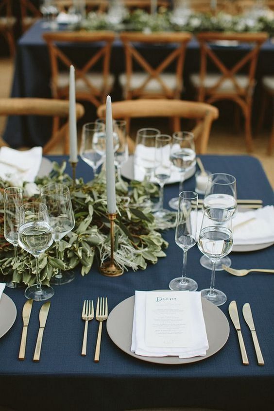 a simple and chic table setting with a navy tablecloth, a lush greenery runner, tall candles and grey plates