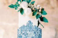 23 a square wedding cake styled as azulejo in blue and white and topped with greenery and neutral blooms