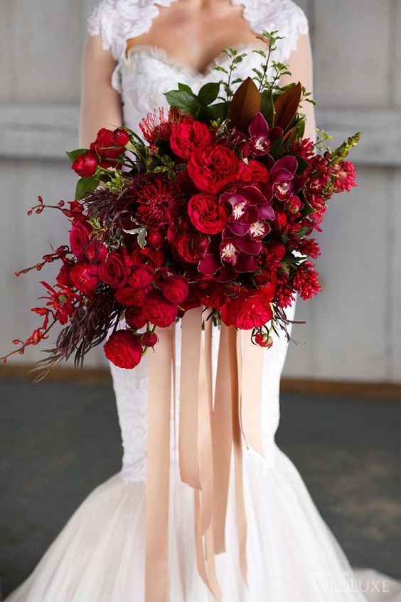 a deep red and fuchsia wedding bouquet with a catchy shape, some blooming branches and touches of foliage plus long ribbons