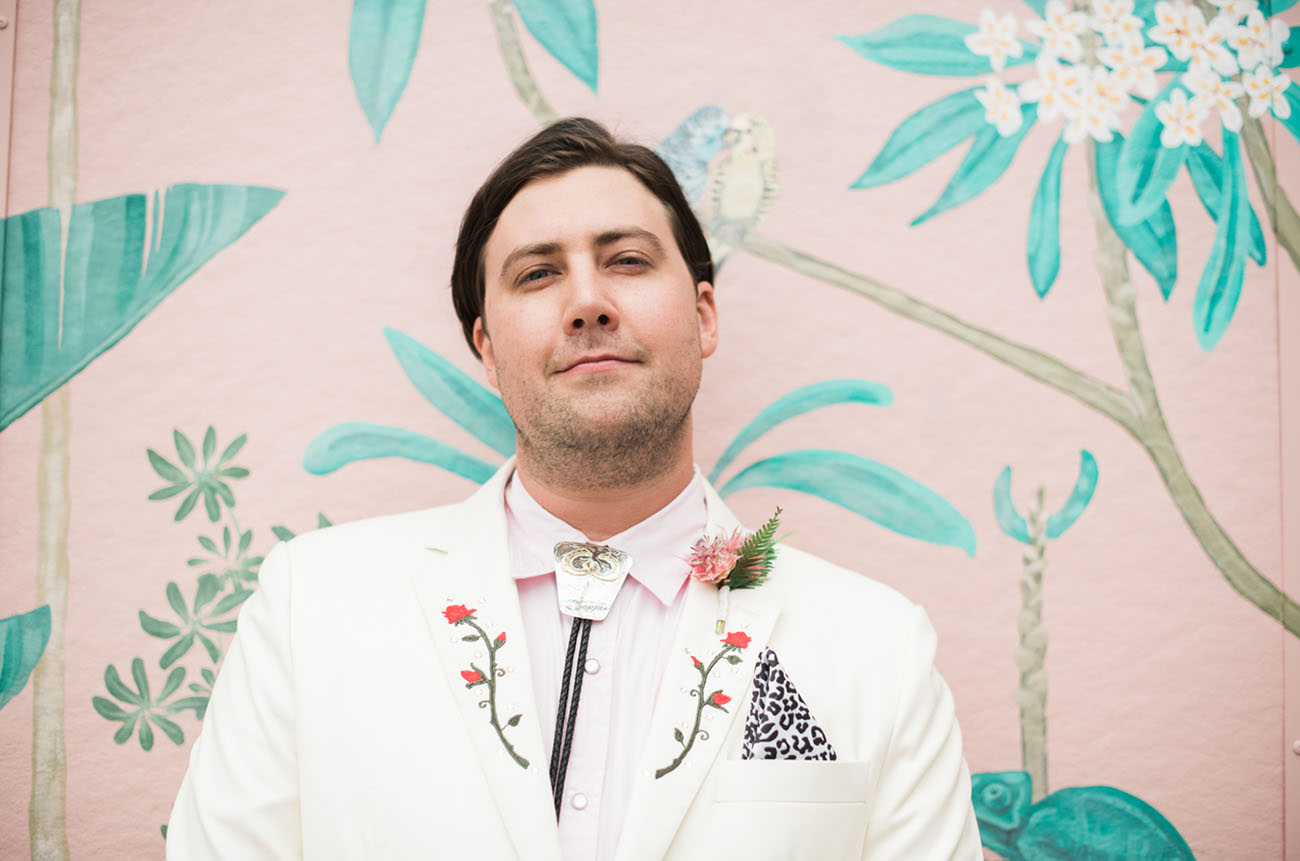 a whimsical groom look with a white suit, a pink shirt, a bolo tie and a leopard handkerchief