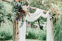 03 a relaxed backyard wedding arch of wood, airy white fabric, colorful blooms and wildflowers and greenery for a boho wedding