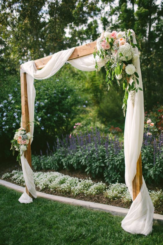 a romantic backyard wedding arch of wood, with airy white fabric and light pink and white blooms and greenery