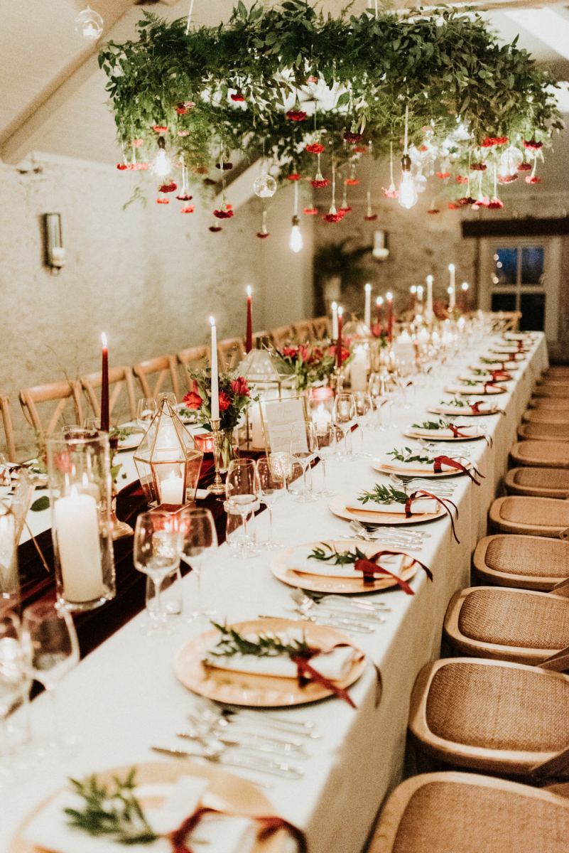The wedding tablescape was done with a burgundy velvet runner, red and white candles, greenery and red blooms and matching chandeliers and wooden chargers