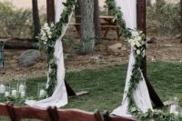 08 a dark stained wedding arch with white curtains, greenery and white blooms, white candles and stained chairs