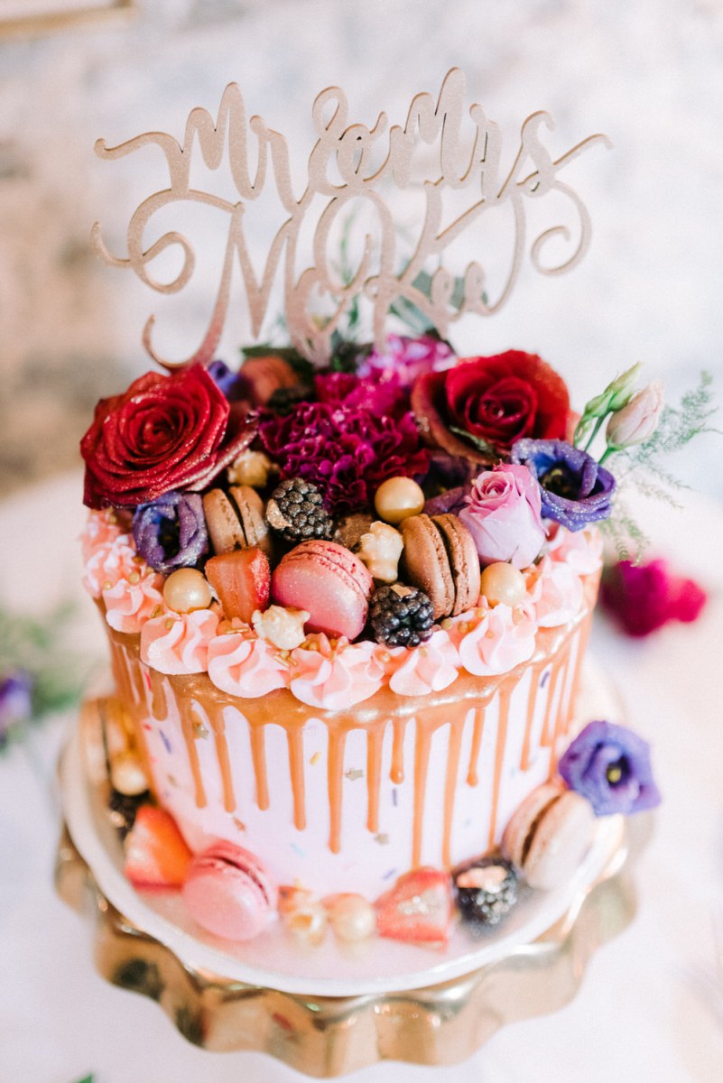 The wedding cake was colorful, pink with chocolate drip, pink and neutral macarons, red and purple blooms, a calligraphy topper and berries