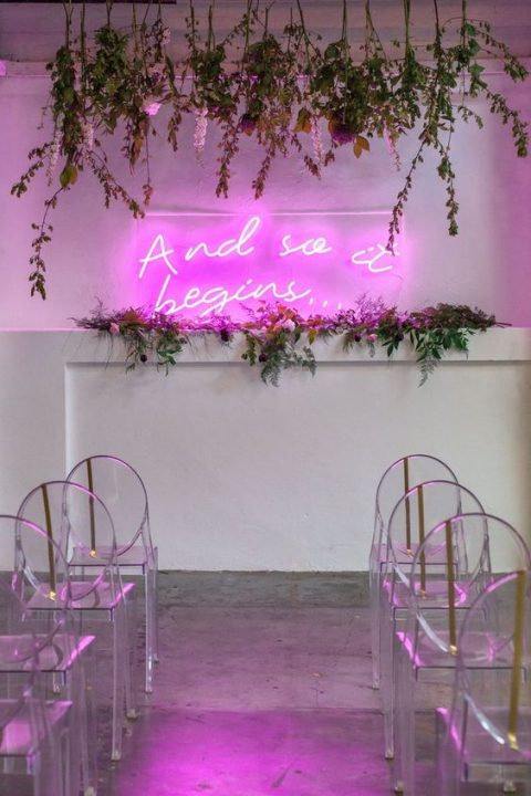 a chic modern wedding ceremony space with a pink neon sign, greenery, hanging blooms and acrylic chairs