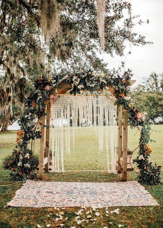 a boho backyard wedding arch with macrame, greenery, bright and white blooms and a boho rug