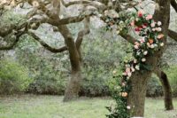 21 a backyard wedding altar of a living tree decorated with greenery, blush and red blooms climbing up it and some simple chairs
