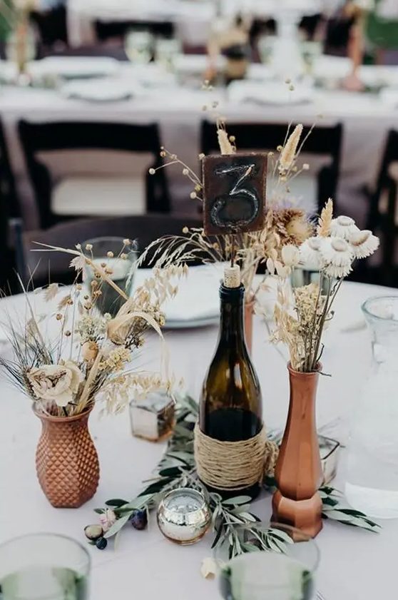 a beautiful rustic wedding centerpiece of rust-colored vases, dried blooms, bunny tails, berries, a candle and olive branches is amazing