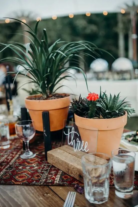 a boho wedding centerpiece with pots with greenery, succulents, a boho table runner and a table number