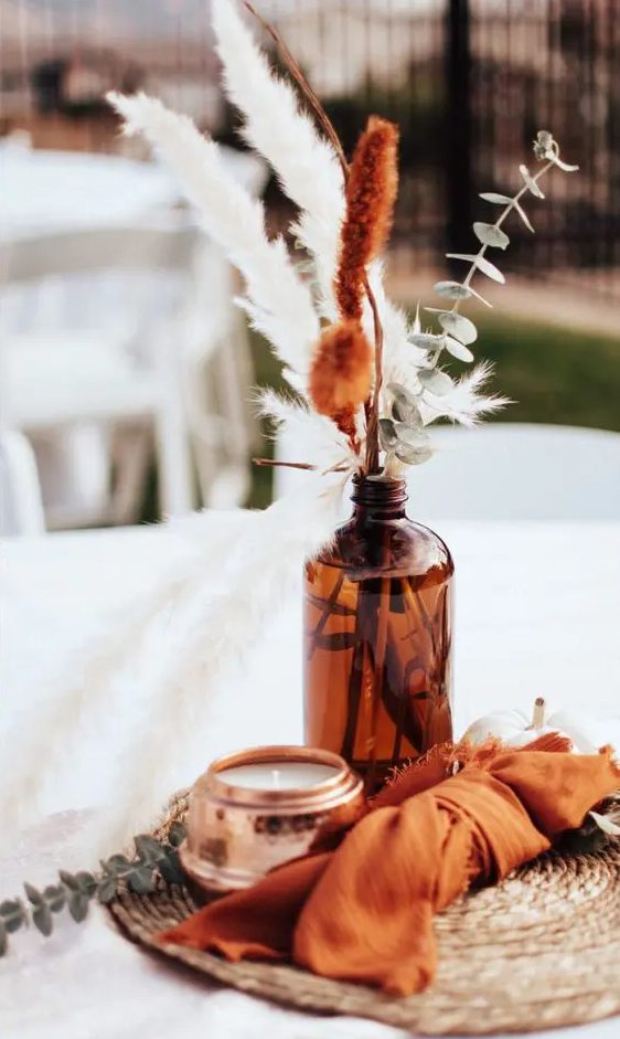 a bright boho wedding centerpiece of an apothecary bottles with greenery and dried grasses, a candle and an orange napkin