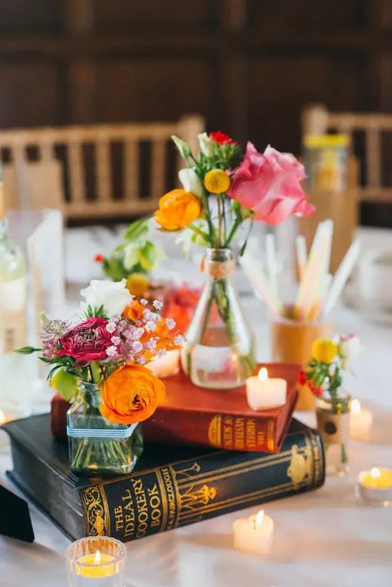 A bright eclectic wedding centerpiece of a book stack, bold blooms and greenery and some small heart shaped candles