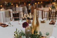 a casual rustic wedding centerpiece of a wood slice, candles, greenery, gilded bottles and burgundy blooms for a backyard celebration