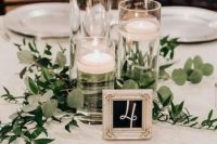 a casual wedding centerpiece of floating candles in tall jars and greenery plus a glam frame table number