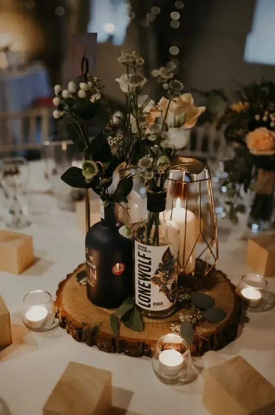 a catchy wedding centerpiece of a wood slice, bottles with greenery, berries and neutral blooms, an elegant candle lantern and candleholders around